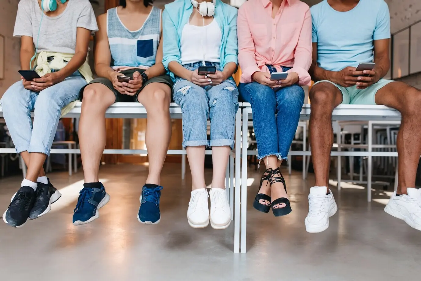 Young man in blue sneakers hanging out with friends, typing a new message on his phone. Indoor portrait of students sitting together on a desk and using smartphones.